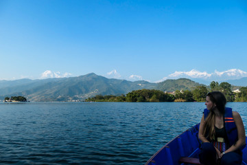 A woman sitting in a blue boat and enjoying a tour across Phewa Lake in Pokhara, Nepal. Behind her there are high, snow capped Himalayas with Mt Fishtail (Machhapuchhare) between them. Relaxation
