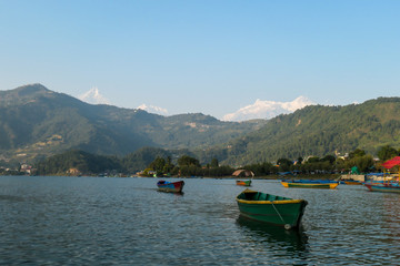 Fototapeta na wymiar A view on Phewa Lake in Pokhara, Nepal with many colorful boats anchored around the shore. There are high Himalayan ranges in the back. Calm surface of the lake. Clear and sunny day. Serenity