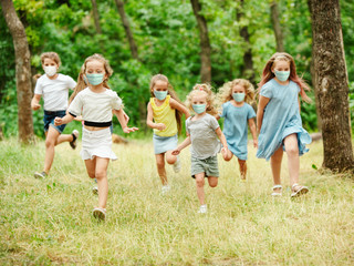 Obraz premium Happy little kids wearing protective face mask jumping and running on meadow, forest. Looks happy, cheerful, sincere. Copyspace. Childhood, pandemic concept. Healthcare, coronavirus pandemic.