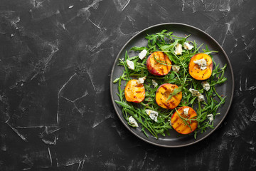 Fresh diet salad grilled peach, arugula and blue cheese in a plate on a black stone background. Top view, flat lay.