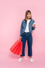 Naklejka premium Woman in jeans and denim jacket using smartphone and holding red shopping bags on pink background