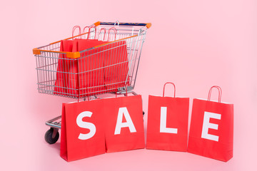 Lettering sale on red shopping bags near cart on pink background