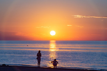 Fototapeta premium Beautiful sunset at the sea. Young people on the beach. Couple are watching the sunset. Sea and red-yellow sky. Silhouettes of people on the background of the sunset. In the evening by the sea