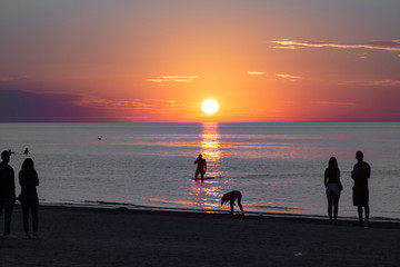 Beautiful sunset at the sea. Young people on the beach. Couple are watching the sunset. Sea and red-yellow sky. Silhouettes of people on the background of the sunset. In the evening by the sea