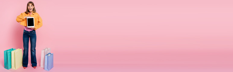 Panoramic concept of excited woman holding digital tablet with blank screen near shopping bags on pink background