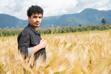 Naklejka na ściany i meble Young hispanic man standing in the middle of a golden wheat field enjoying the sunset in the summer - young dark-haired man taking off his jacket surrounded by mountains