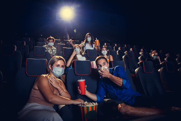 Cinema, movie theatre during quarantine. Coronavirus pandemic safety rules, social distance during movie watching. Men and women wearing protective face mask sitting in a rows of auditorium, eating