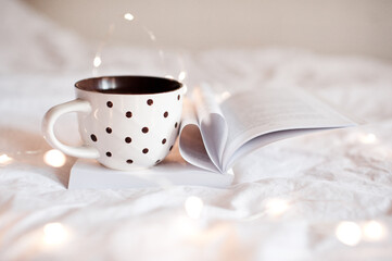 Fototapeta na wymiar Cup of tea on open book with heart shape folded pages closeup in bed at night. Good morning.