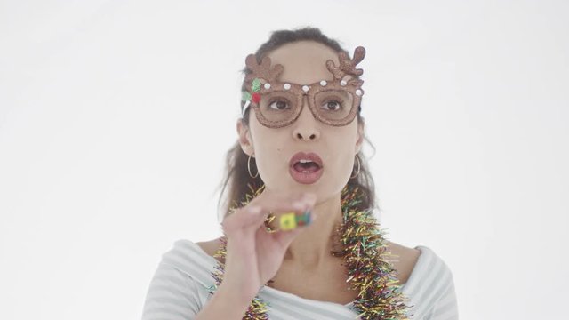 4K portrait of beautiful young brunette with ponytail and Christmas accessories in festive mood blowing in multi-coloured party hord. 360 degree tracking arc shot movement.
