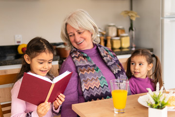 Cheerful young granddaughter with book reading stories for her multigenerational family at kitchen table, inside. Relationship, leisure, enjoyment, aging concept.