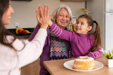 Obraz na płótnie Canvas Cheerful brazilian family of women with arms raised giving a high five in kitchen home, indoors. Unity, happiness, affection, love, care concept.