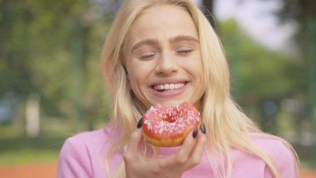 Portrait of cheerful blond woman biting sweet donut and laughing. Happy beautiful Caucasian lady eating delicious pastry outdoors on sunny summer day.
