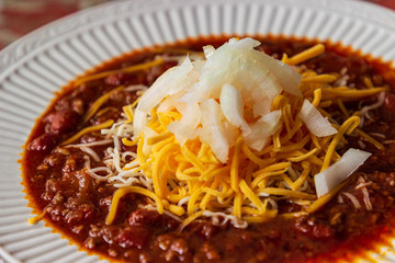 A bowl of delicious homemade chili in a white bowl and garnished with cheddar and Monterrey Jack cheeses and diced onions.