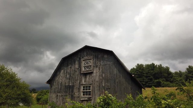 Scenic old rustic farm building in rural countryside farmland by green grass, vegetation and trees, pan up to grey stormy blanket of clouds in cloudy sky, handheld
