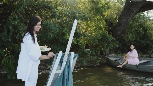 Young painter paints a real landscape on the textile, a girl sitting in a boat on the shore of a lake.