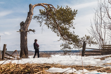 A man stands next to a crooked pine tree in a spring field