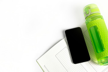 Training diary or meal plan, mobile phone and green fitness bottle laying on plain white background. Mobile sports application concept for measuring calories.