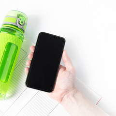 Green fitness bottle, training diary and black mobile phone mock up in arm. Training application concept. Square format banner for social media.