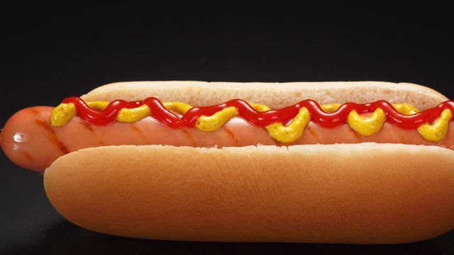 Hot dog with grilled sausage, ketchup and mustard on a black background.
