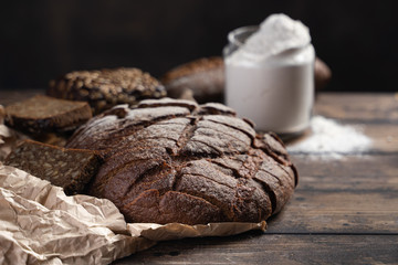 Whole loaf and pieces of homemade rye bread placed on paper on lumber rustic table near flour