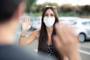 Young beautiful woman student wearing surgical mask and waving hand for greeting to her friend in...