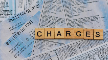 Lettres en bois composant le mot "charges" sur un fond de bulletins de paie français. Concept de cotisations sociales élevées