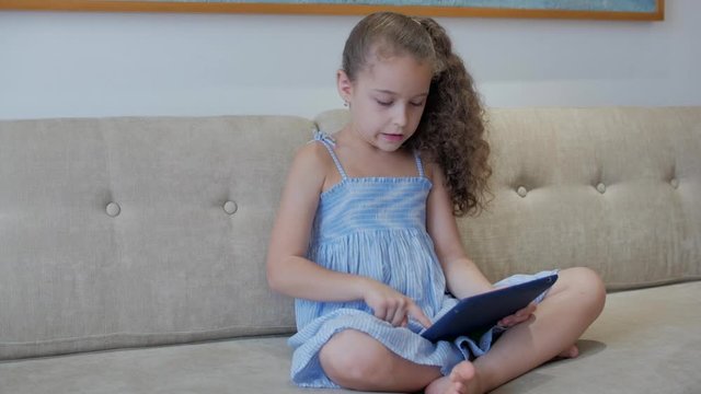 Cute little girl preschool child using a digital tablet technology device looks telephone.Plays in the phone sitting on the sofa alone.Children tech addiction concept play game at home.