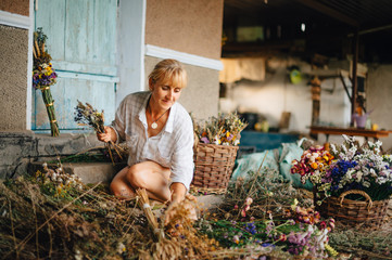 Naklejka na ściany i meble Positive woman in a shirt at the cottage creates bouquets of dried flowers with a smile on his face. Amateur florist makes compositions from flowers in the country.