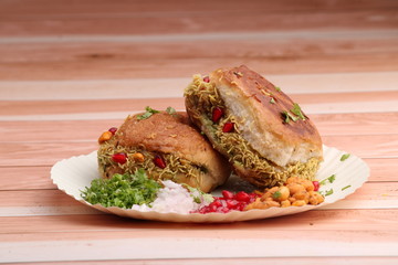 Dabeli is an Indian snack item served with Pomegranate Seeds and Cilantro in white paper plate. over wooden background