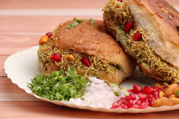 Dabeli is an Indian snack item served with Pomegranate Seeds and Cilantro in white paper plate. over wooden background
