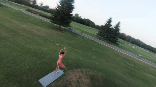 Creative aerial shot with fpv racing drone of woman in red tracksuit doing yoga in the park at sunny day at sunset.