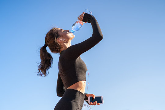 Thirsty Young Fit Woman Drinking Water From Plastic Bottle, Resting After Jogging, Holding Mobile Smartphone In Her Hand, Listens To Music. Blue Sky With Copy Space. Hot Day. 