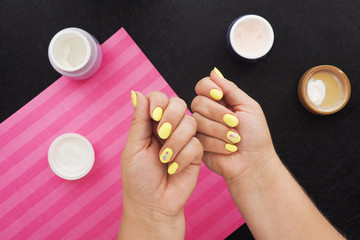 female hands with bright yellow manicure. on the black table there are jars of cream, varnish and a pink napkin..