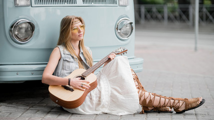 A hippie woman sits on the sidewalk and plays guitar next to a classic minivan.