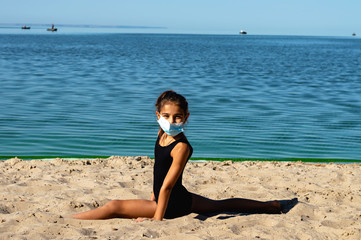 A gymnast girl in a protective medical mask, dressed in a sports swimsuit, performs gymnastic exercises on the beach. Isolation from people. Covid virus 19. Pandemic disease, quarantine, outdoor sport