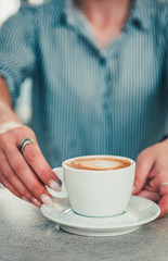 Closeup female barista hands holding cup of aromatic latte or cappuccino coffee in bar or cafe