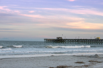 The Apache Pier at sunset in Myrtle Beach South Carolina. Waves are at the foreground with blues and pinks in the sky.