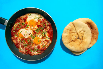 Shakshuka - poached eggs in tomato sauce, onion, pepper and spices in iron pan with pita bread on blue bright background. Famous traditional Arabic and Israeli breakfast - chakchouka with flatbread