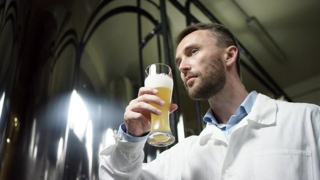 A male laboratory worker in a white robe is degusting a first set of a beer. A bottom view slow motion close up