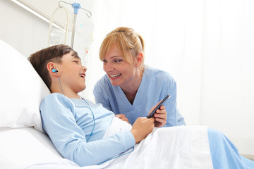 Obraz premium Happy child lying in bed in hospital room and smiling nurse using smartphone surfs the internet wearing earphones