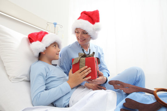 Christmas Holiday In Hospital Happy Child Lying In Bed With Santa Claus Hat And Nurse Giving Him A Present