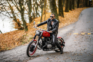 Fototapeta na wymiar Bearded brutal man in sunglasses and leather jacket sitting on a motorcycle on the road in the forest
