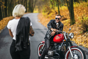 Naklejka na ściany i meble Pretty couple near red motorcycle on the road in the forest with colorful blured background