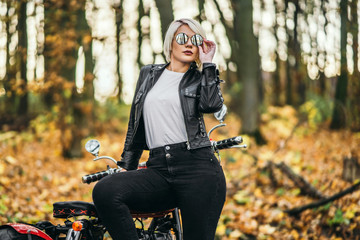 Pretty blonde biker girl in sunglasses with red motorcycle on the road in the forest