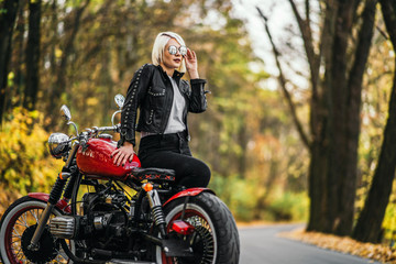 Fototapeta na wymiar Pretty blonde biker girl in sunglasses with red motorcycle on the road in the forest