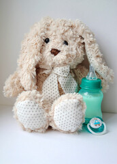 Soft baby toy rabbit next to a baby bottle with milk and a pacifier on a white background. The concept of childhood and motherhood.