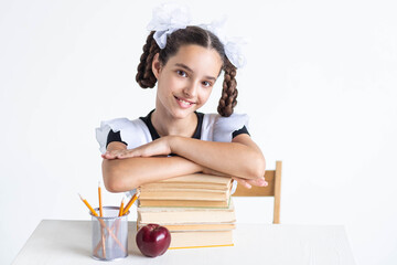 Back to school concept.Close up portrait of smiling schoolgirl girl in school uniform sitting at the table with her hands on a stack of books near pencils and a red apple on a white wall background.
