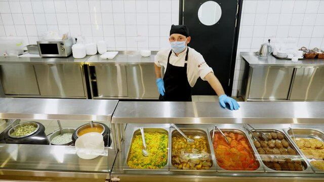 a cook or waiter, in protective gloves and mask, stands behind display case with different dishes, in self-service cafeteria or buffet restaurant. health food. volunteering and charity. reopening