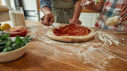 Cropped shot of couple making pizza together at home. Man in apron adding, applying tomato sauce on the dough while woman adding mozzarella cheese. Hobby, lifestyle