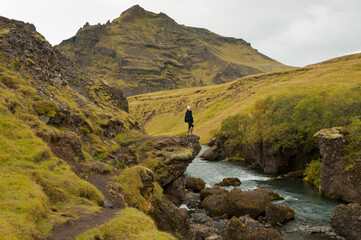 A girl wearing a hat and wellington boots stands on the edge of a rock on top of a river while contemplating the stunning Icelandic landscape